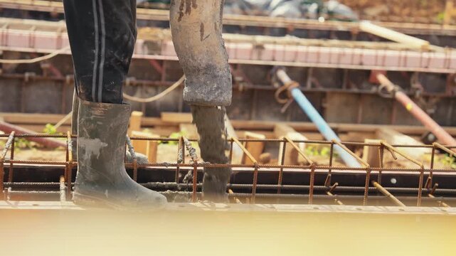 Construction worker in rubber boots pouring wet concrete from a flexible hose into a rebar reinforced formwork, creating a solid building foundation at a construction site in warm sunlight