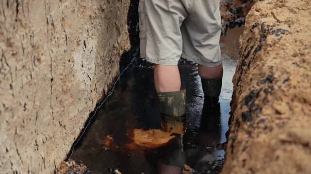 Worker wearing rubber boots carefully walking through a narrow, muddy trench filled with dark, dirty water, inspecting the area for potential damage or contamination