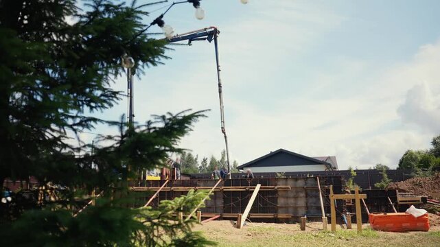 Industrial machinery with a long boom arm pouring liquid concrete into a house wall formwork on a sunny day. Construction workers are managing the process of building a new modern home foundation