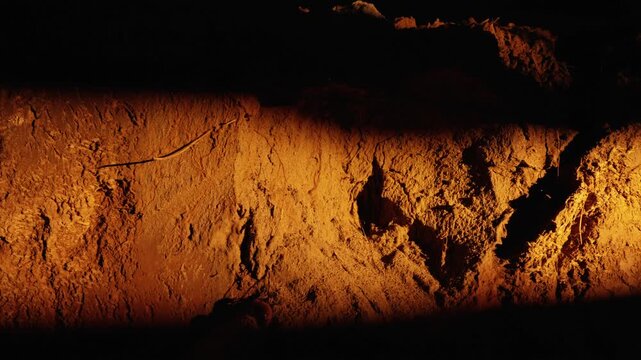 Warm light from a headlamp or flashlight moving across the rough, textured surface of an ancient cave wall, revealing crevices, cracks, and details of the rocky underground passage