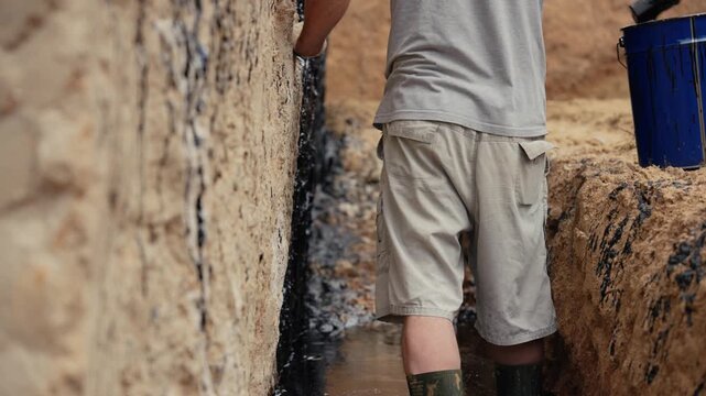 Male worker standing in a muddy trench applying black bitumen mastic with a trowel to the concrete foundation of a house for damp proofing and waterproofing protection on a construction site