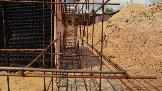 Close up view moving through a reinforced steel cage in a trench, prepared for pouring concrete to create a monolithic strip foundation for a residential building at a construction site