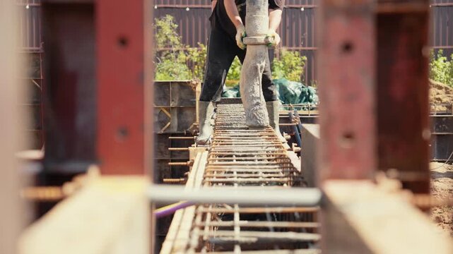 Construction worker in protective gear pouring wet cement from a pump hose into a reinforced steel rebar cage, creating a solid foundation for a new building during a sunny day
