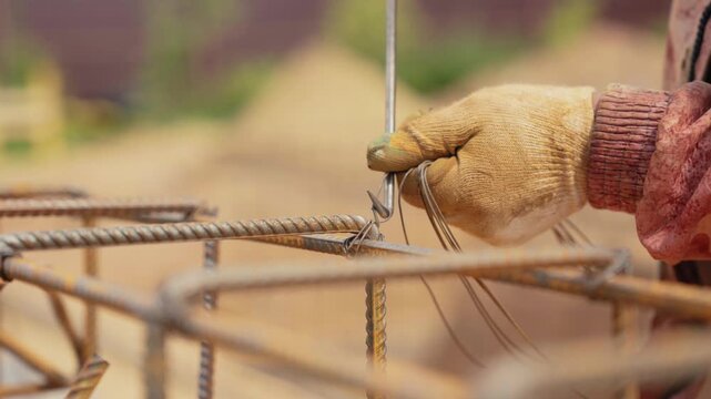 Closeup view of a construction worker's gloved hands skillfully using a tying tool to secure steel rebar with wire, preparing the reinforcement cage for a concrete foundation on a building site