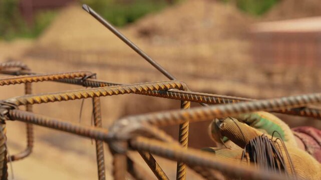 Close up of a construction worker's hands in gloves using a hook tool to tie steel rebar with wire, preparing the reinforcing cage for pouring a concrete foundation at a building site