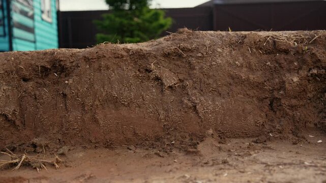 Close up of a construction excavator bucket scooping up dark earth from the ground during excavation work on a building site, with a blurred background showing a fence and a house