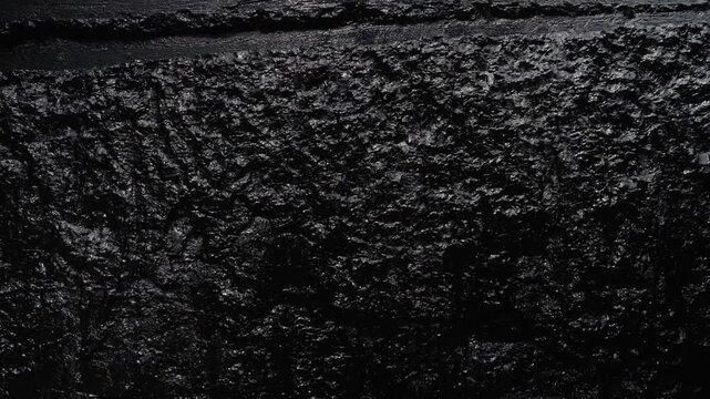 Extreme close up of a dark, rugged stone surface with a coarse texture and natural irregularities. This detailed view reveals the intricate patterns and shadows of the raw material
