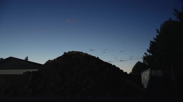 Silhouettes of a backyard landscape with a large mound of dirt against a darkening twilight sky. The serene scene captures the transition from dusk to night in a quiet suburban setting