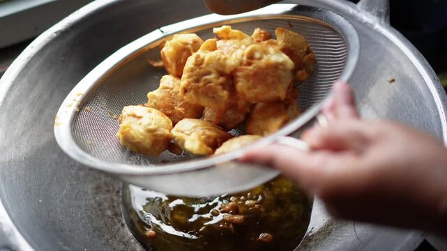 Hand removing golden fried food items from hot oil using a metal strainer over a large pot. Steam rises as excess oil drains. Close-up shot of cooking process with focus on texture and color.