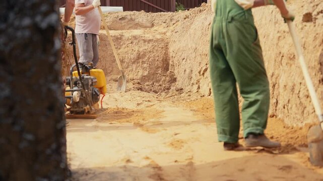 Professional builders compacting sand at the bottom of a foundation pit with a vibrating plate compactor. A colleague is digging nearby with a shovel during excavation work on a construction site
