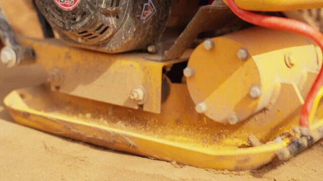 Construction site close up showing a vibratory plate compactor machine working on compacting sand, with sand grains bouncing from the powerful ground vibration during the soil tamping process