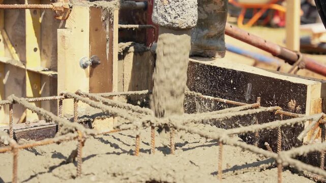 Liquid cement mixture being poured from a pump hose into a wooden formwork with steel rebar cage, creating a solid foundation for a new building during a sunny day on a construction site