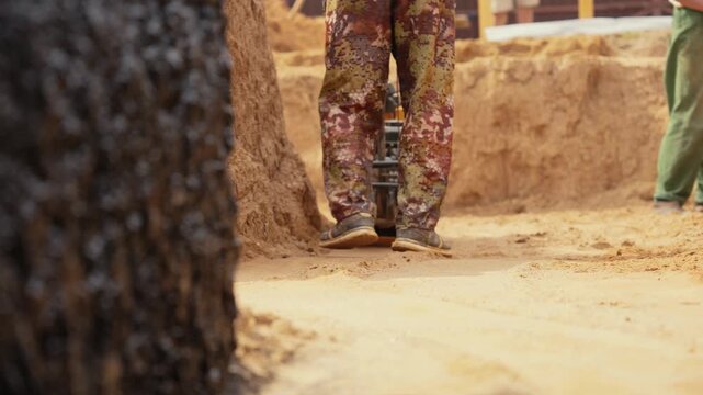 Road construction worker in camouflage pants using a vibratory tamping rammer to compact the sandy ground for a new foundation, with another worker visible in the background with a shovel