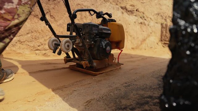 Close up view of a construction worker using a vibratory plate compactor to level and compact sand at a building site, preparing the ground foundation for laying paving or asphalt
