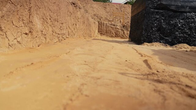 Camera movement along a trench foundation with walls waterproofed with black bitumen mastic, showcasing the initial stages of building construction on a bright, sunny day at a new development site