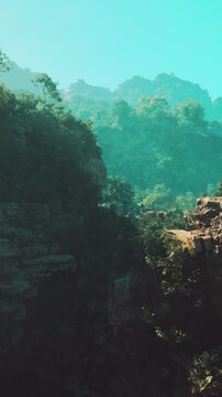 A view of rocky formations surrounded by greenery under a clear blue sky. Trees are scattered throughout the landscape. The sun shines brightly on the mountains.