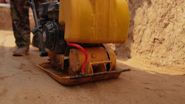 Construction worker operating a vibratory plate compactor to level and compact sand, creating a solid foundation for paving during roadworks, with dust and grains flying from the vibration