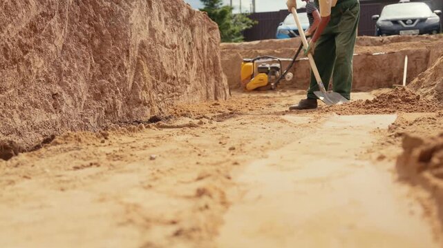 Professional construction worker in overalls using a shovel to level sand at the bottom of a foundation trench during the initial stages of building a house at a residential construction site