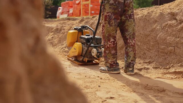 Construction worker in camouflage pants tamping sand with a vibrating plate compactor machine, preparing the foundation and leveling the ground at a building site during daytime earthworks