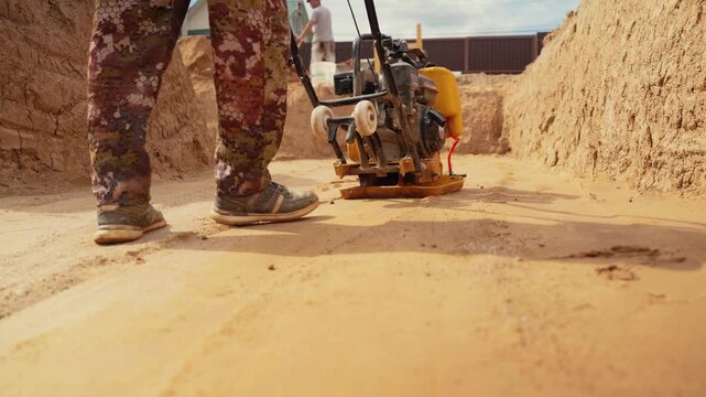 Construction worker operating a vibratory plate compactor to level and compact sand at a construction site. Powerful machinery is used for soil compaction and ground preparation work for a foundation
