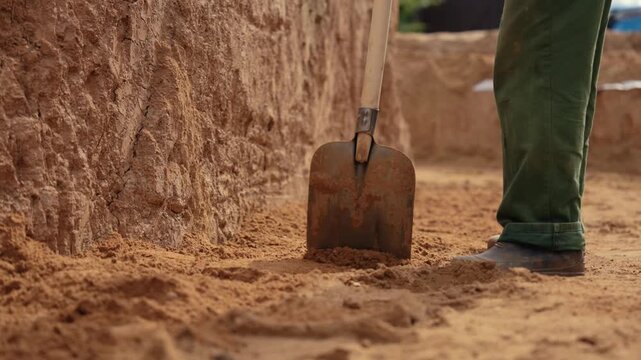 Close up view of a construction worker using a shovel to move sand and dirt at a building site, showing the boots and shovel in detail as part of the manual labor for site preparation