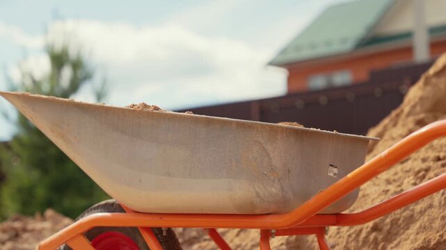 Close up of a worker shoveling sand into a wheelbarrow at a construction site, with coarse sand particles falling into the cart during building foundation work in slow motion