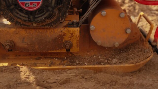Construction equipment working on a site, with a vibrating plate compactor machine leveling and tamping loose sandy soil for a solid foundation during groundwork preparation