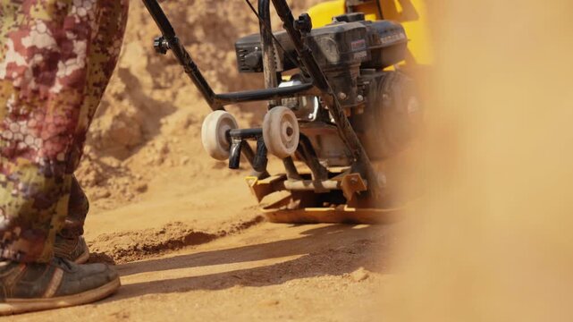Close up view of a builder operating a vibratory plate compactor, leveling the sand ground at a construction site and raising clouds of dust in slow motion on a sunny day