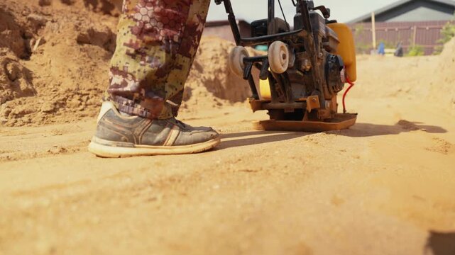 Professional construction worker using a vibratory plate compactor machine to level and compact sand at a building site, preparing the foundation for paving or construction projects