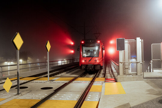 A vibrant red train approaches an illuminated station in dense fog, symbolizing movement and transition against the backdrop of a mysterious urban night.