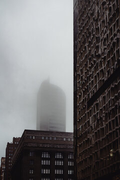 The peak of a tall building is obscured by a thick layer of fog, creating an enigmatic atmosphere that elicits feelings of curiosity and wonder about the urban landscape.