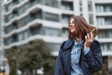 Fototapeta premium Young adult woman with brunette hair wearing a denim shirt and casual jacket talks on her smartphone in an urban street scene, smiling as modern architecture frames the shot.