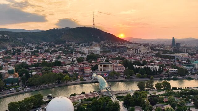 Panoramic aerial drone view of Tbilisi cityscape at golden hour sunset, showing the iconic glass Bridge of Peace over Kura River, with Mount Mtatsminda and TV tower rising in the background, Georgia