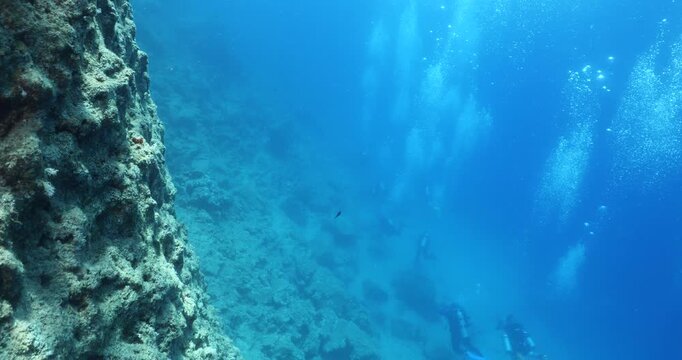  scuba divers exploring around a reef underwater deep blue water big rocks and bubbles ocean scenery 
