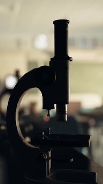 A microscope stands on a table in a classroom. Students are engaged in a science lesson. The room has a soft focus background. Light enters through the windows.