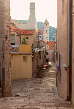 Narrow stone street with historic buildings, outdoor cafe tables and a bell tower in Dubrovnik Old Town, Croatia.