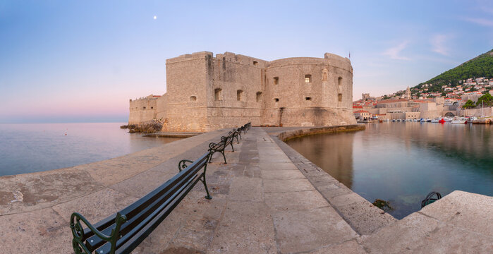 Stone fortress and harbor pier at dawn in Dubrovnik, Croatia, with calm Adriatic Sea and early morning light.