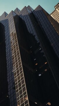 A tall building covered in glass stands in an urban area. The structure reflects sunlight during the day. Other buildings are visible in the background along with a blue sky.