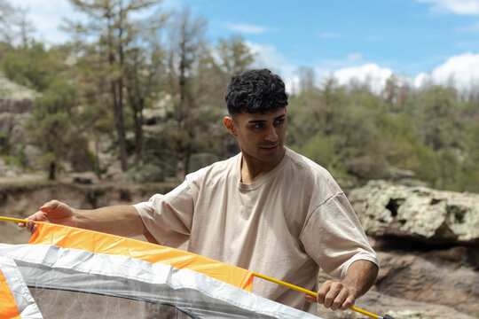 Young man actively setting up an orange camping tent while enjoying an outdoor lifestyle, preparing for adventure on a sunny day in the mountains