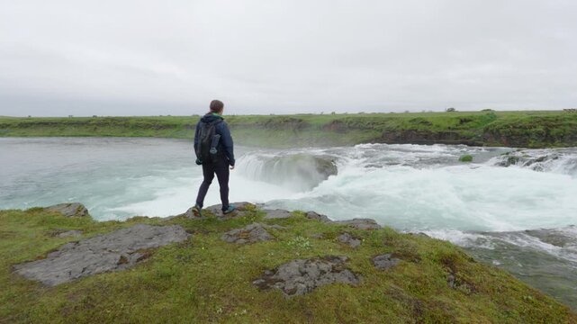 A hiker stands by the powerful &AElig;giss&iacute;&eth;ufoss waterfall on Iceland, surrounded by rugged green landscape and a cloudy sky. The glacial river cascades, creating white foam.