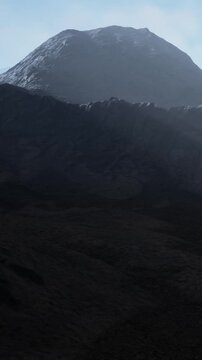 Mountains stand tall in the background while the ground is dark and rough. A mist surrounds the peaks and the sky shows a soft light. The scene is filled with natures raw beauty.