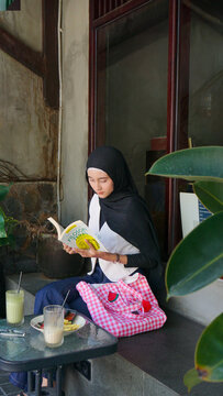 Young Asian Muslim Woman in Black Hijab Reading Filosofi Teras Book Sitting at Outdoor Cafe Table