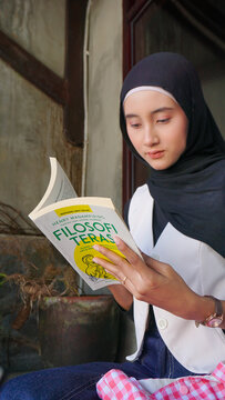 Young Asian Muslim Woman in Black Hijab Reading Filosofi Teras Book Sitting at Outdoor Cafe Table