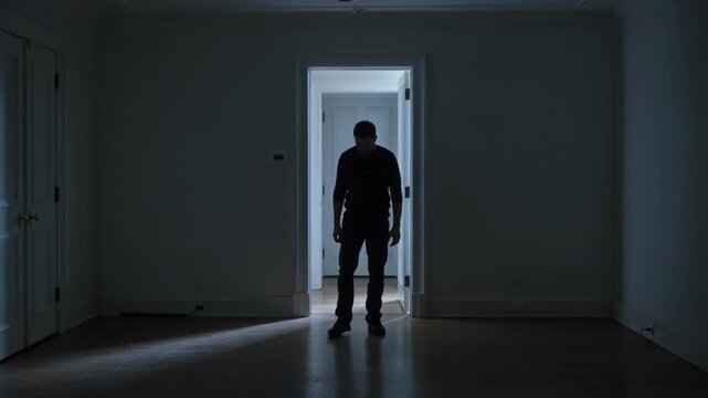 Dark silhouette of a man standing in a doorway backlit with dim cold light. Lonely figure waiting in a dark room. Mystery, solitude and cinematic mood concept