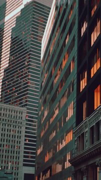 Bright windows reflect the colors of the evening sky in a busy city. Tall buildings rise towards the sky as sunlight fades. People move around the streets below.