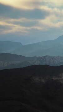 A range of mountains with varying heights and dark shades. Clouds hover above, providing an atmosphere during the evening. The landscape appears rugged and vast.