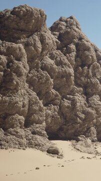 A group of large rock formations rises from sandy terrain in a desert setting. The clear blue sky provides bright light, enhancing the natural colors. Footprints lead towards the rocks.