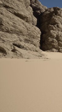 Rocks stand tall near the shoreline. The sand stretches along the beach. The sun shines bright in the clear sky. Nobody is present in this natural scene.