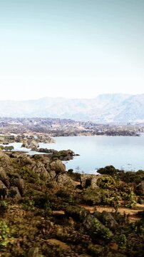 A wide view shows a calm lake bordered by hills and rocks. There are trees and open land visible around the water. The sky is clear and blue, making the scene bright.
