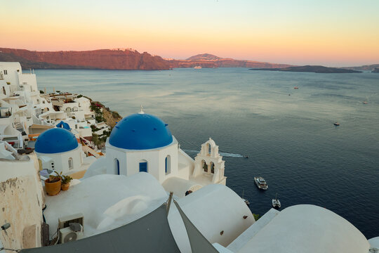 The blue‑domed church of Oia overlooks the Aegean Sea at sunset in Santorini, Greece, glowing in soft Mediterranean light above the tranquil caldera.